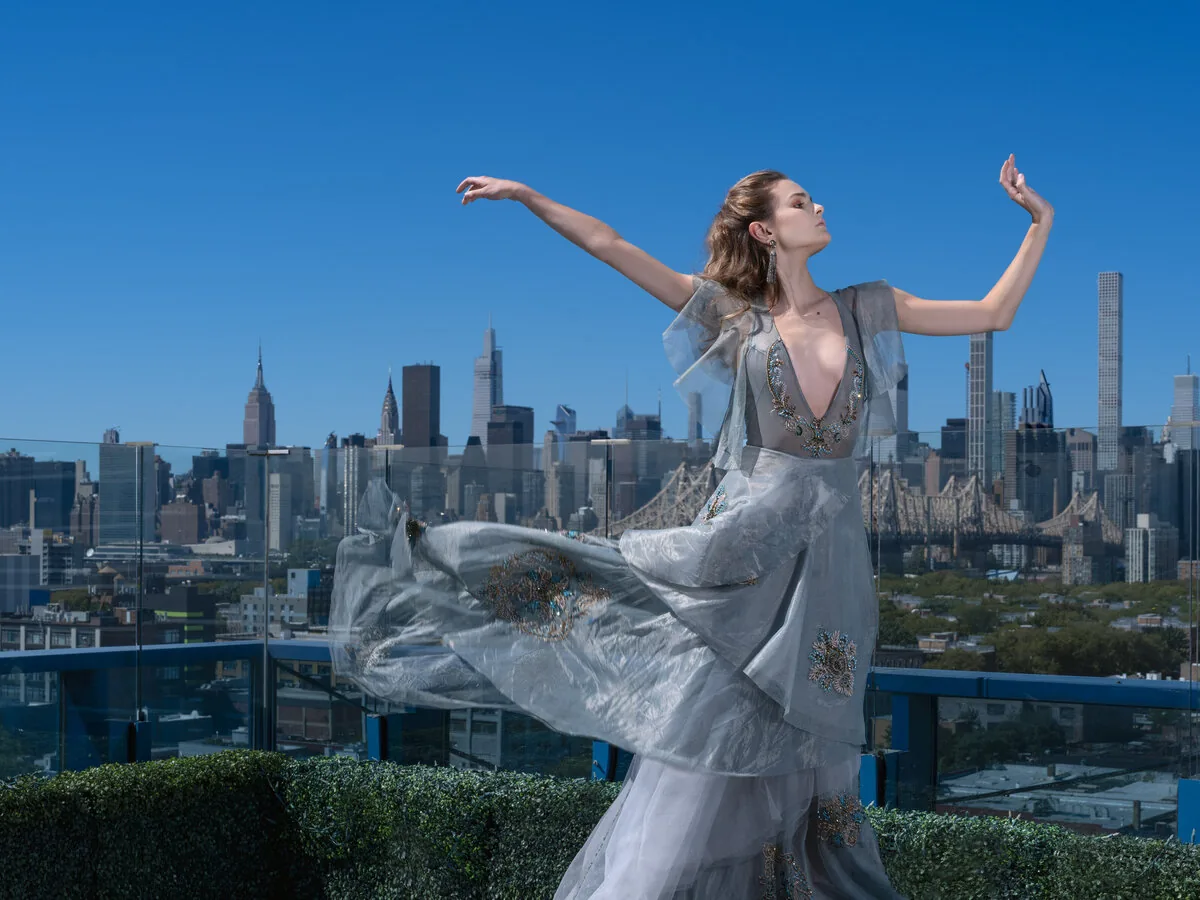 A dancer in a beaded gown extends her arms on a Manhattan rooftop, the skyline behind her.