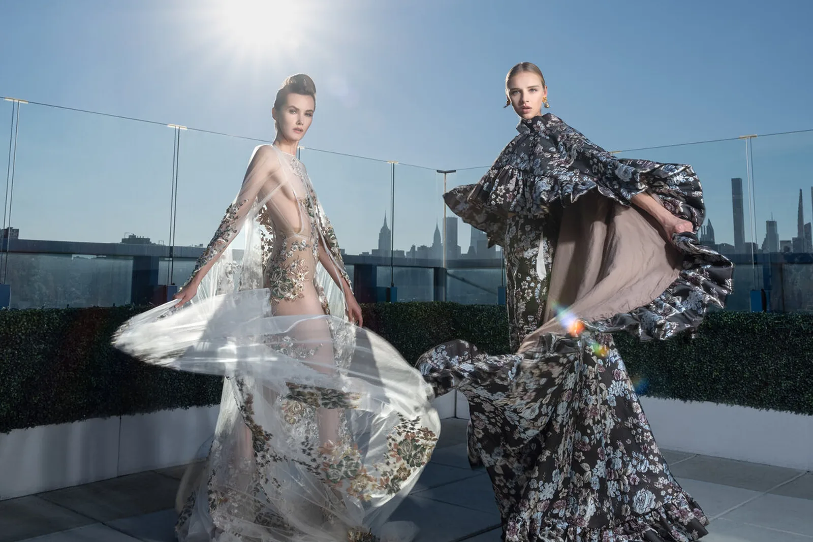 Two models in couture gowns on a Manhattan rooftop, skirts caught in the wind against the skyline.